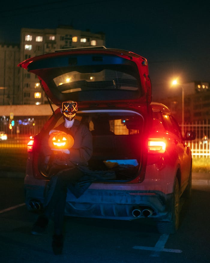 services-04 Urban Halloween scene with a glowing pumpkin and masked person sitting in a car trunk at night.