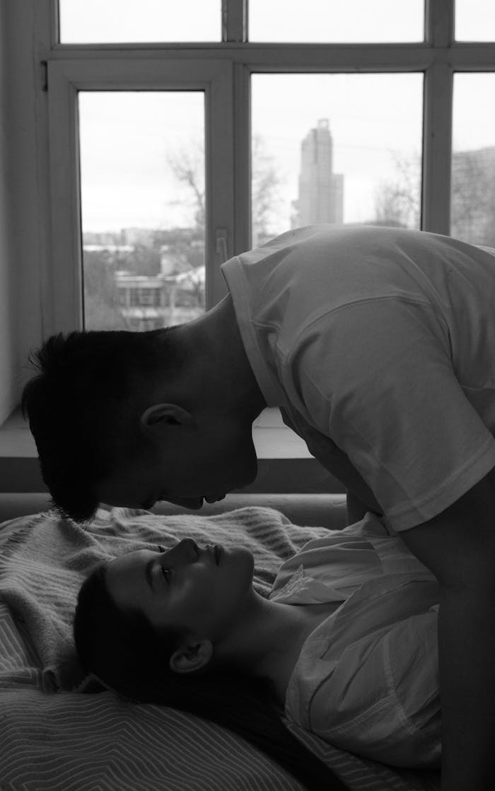 A black and white photo of a couple embracing intimately in a cozy bedroom setting.