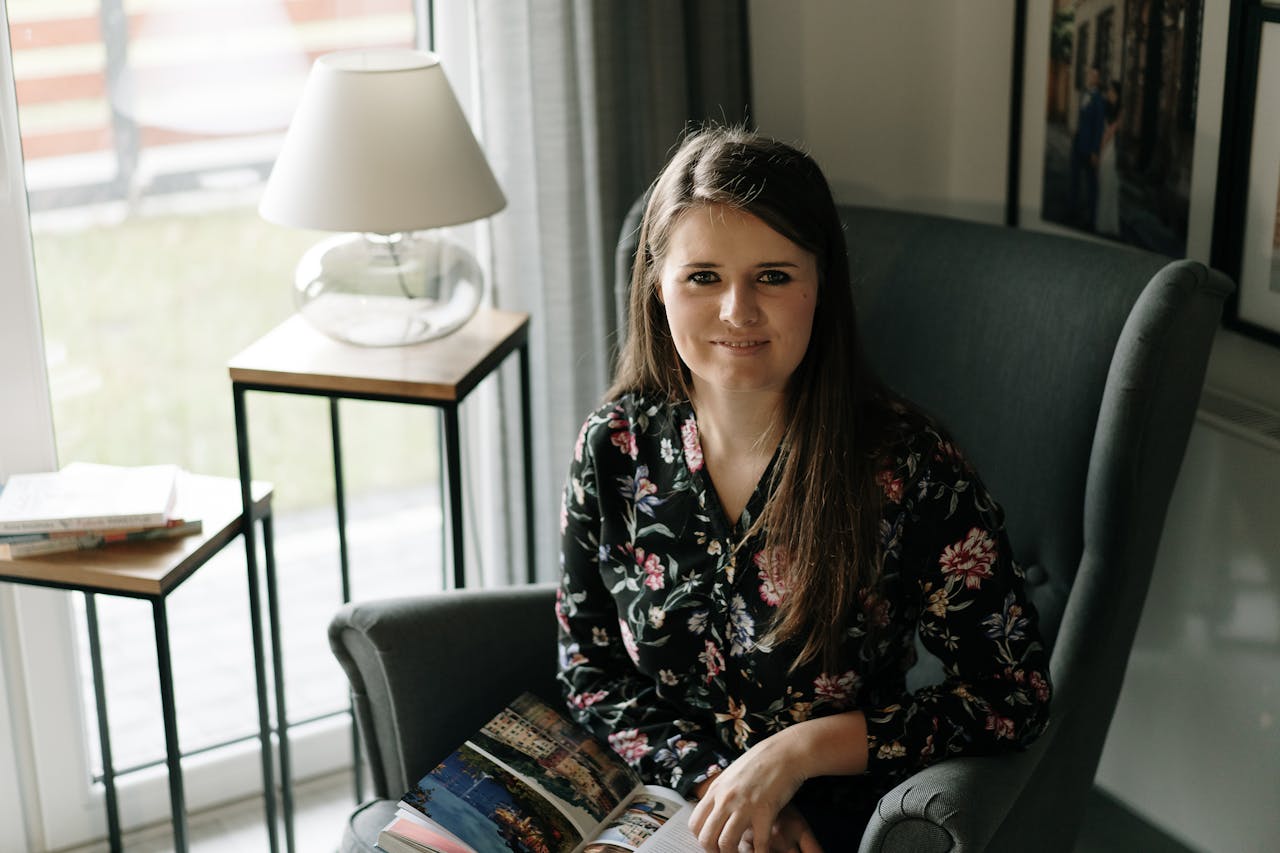 Brunette woman sitting comfortably in an armchair, enjoying a book in a cozy, well-lit living room.